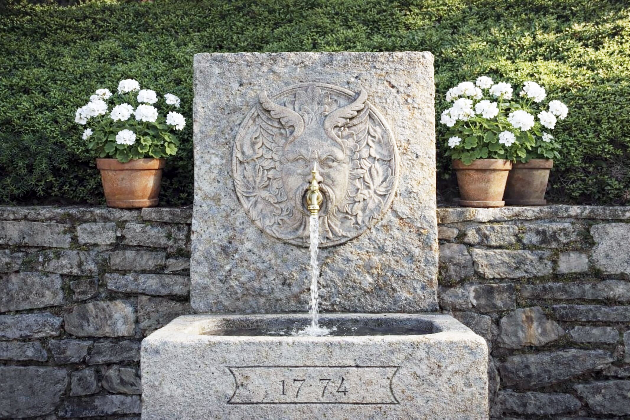 Eine Steinfountain mit Wasserhahn, umgeben von Kübeln mit weißen Blumen, und ein Datum auf dem Sockel.