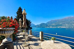 An outdoor terrace with tables and chairs, a stone planter with red flowers, and a view of a lake with mountains in the background. - Villa Marina - Como Lake (Bellano)