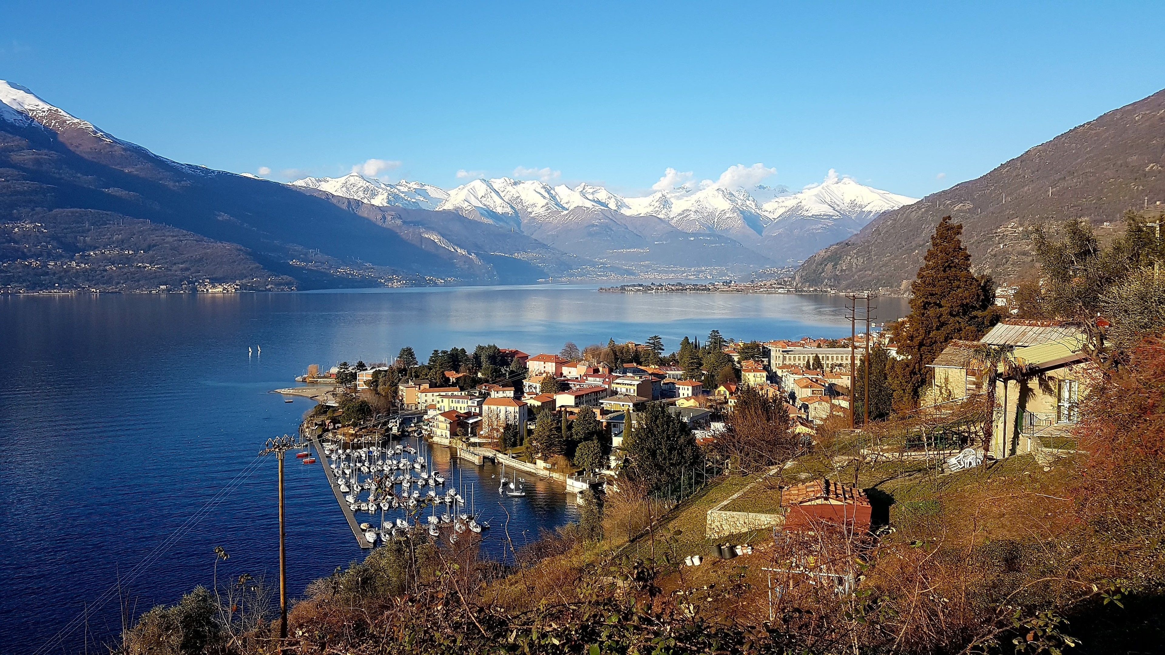 Un pueblo a orillas de un lago con un puerto deportivo, edificios y montañas nevadas al fondo.