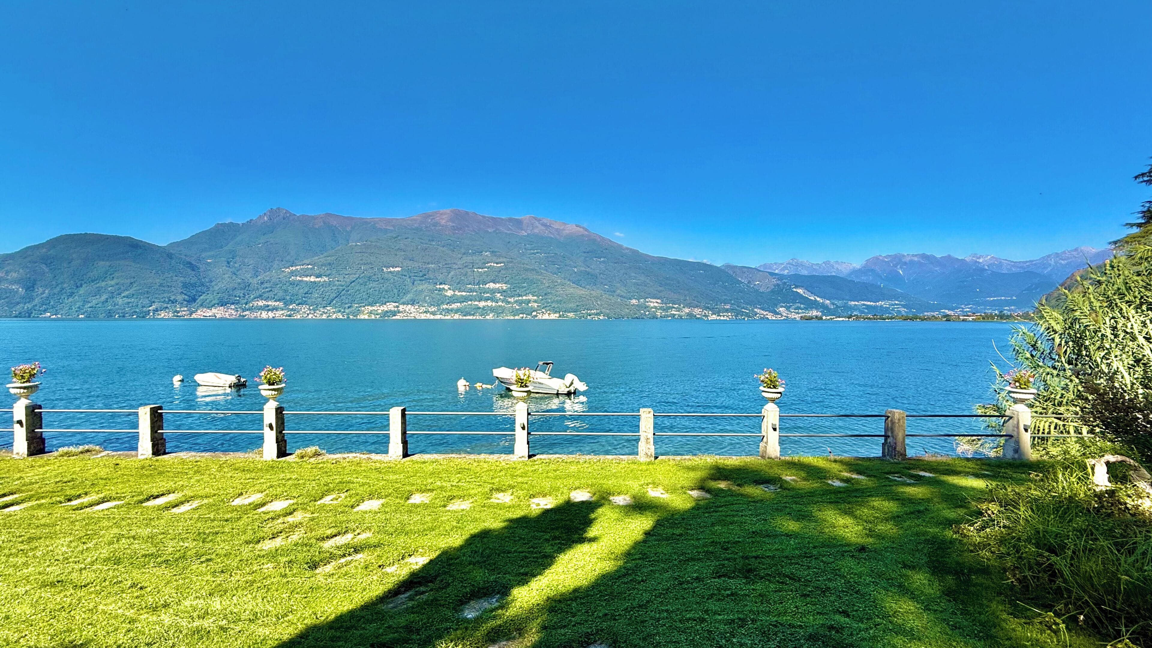 Blick auf einen See mit Booten, einer grünen Wiese und Bergen im Hintergrund.