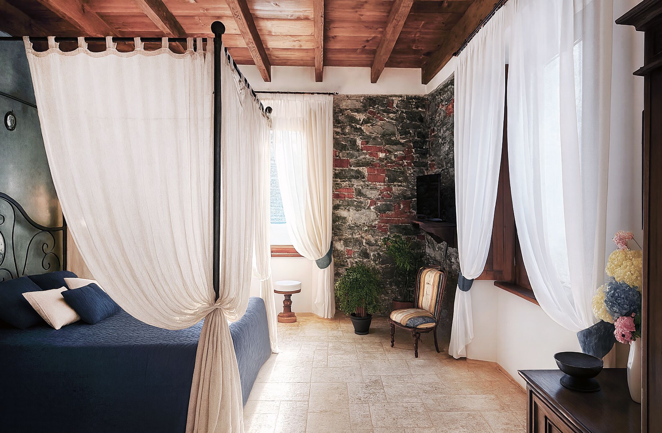 Une chambre à coucher avec un lit à baldaquin, un mur en pierre, une télévision, une plante en pot, une chaise et une petite table.