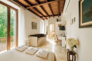 A bedroom with a bed, a sofa, a desk, and a TV. There are towels on the bed and a vase with flowers on a table. - Villa Marina - Como Lake (Bellano)