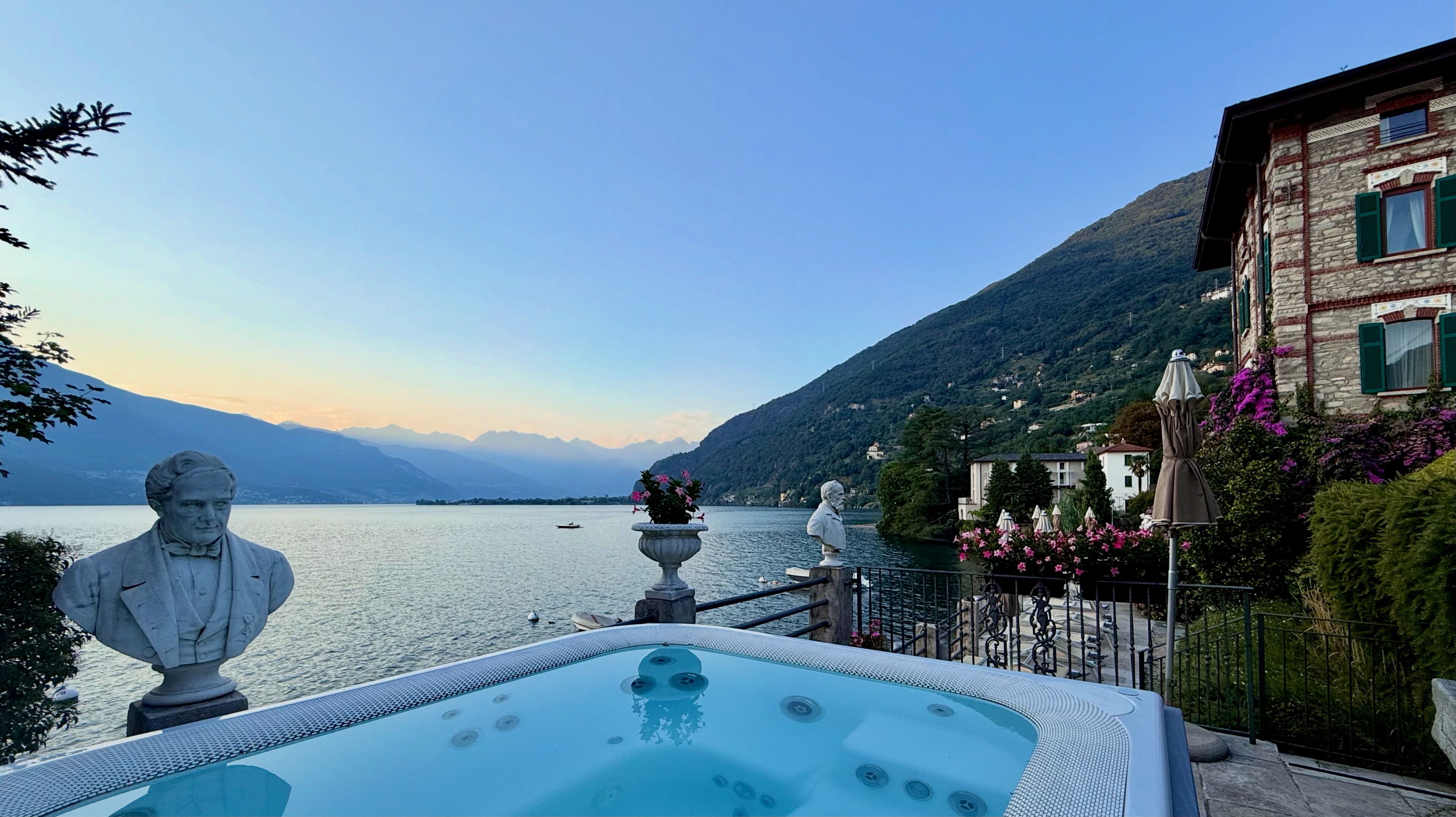 A hot tub with a view of a lake and mountains, a statue, and a building with a balcony.
