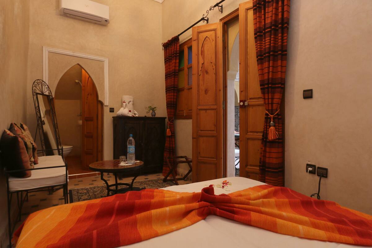 A hotel room with a bed, a table with a bottle and glasses, a mirror, and a wooden wardrobe.