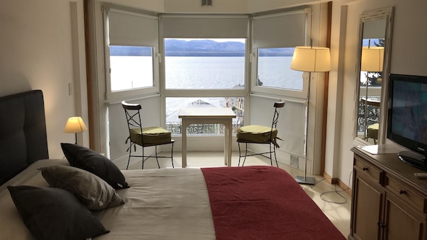 A bedroom with a bed, two chairs, a TV, and a balcony with a view of the sea. - Salta Lake View (San Carlos de Bariloche)
