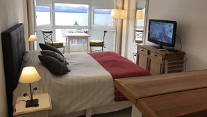 A hotel room with a bed, a TV, a desk, a chair, a table, and a view of a body of water through large windows. - Salta Lake View (San Carlos de Bariloche)