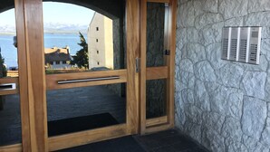A wooden door with a glass panel, a stone wall, and a view of a building and water. - Salta Lake View (San Carlos de Bariloche)