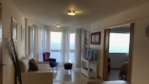 A spacious living room with a sofa, a chair, a wooden table, and a TV on a stand. Large windows offer a view of the sea. - Salta Lake View (San Carlos de Bariloche)