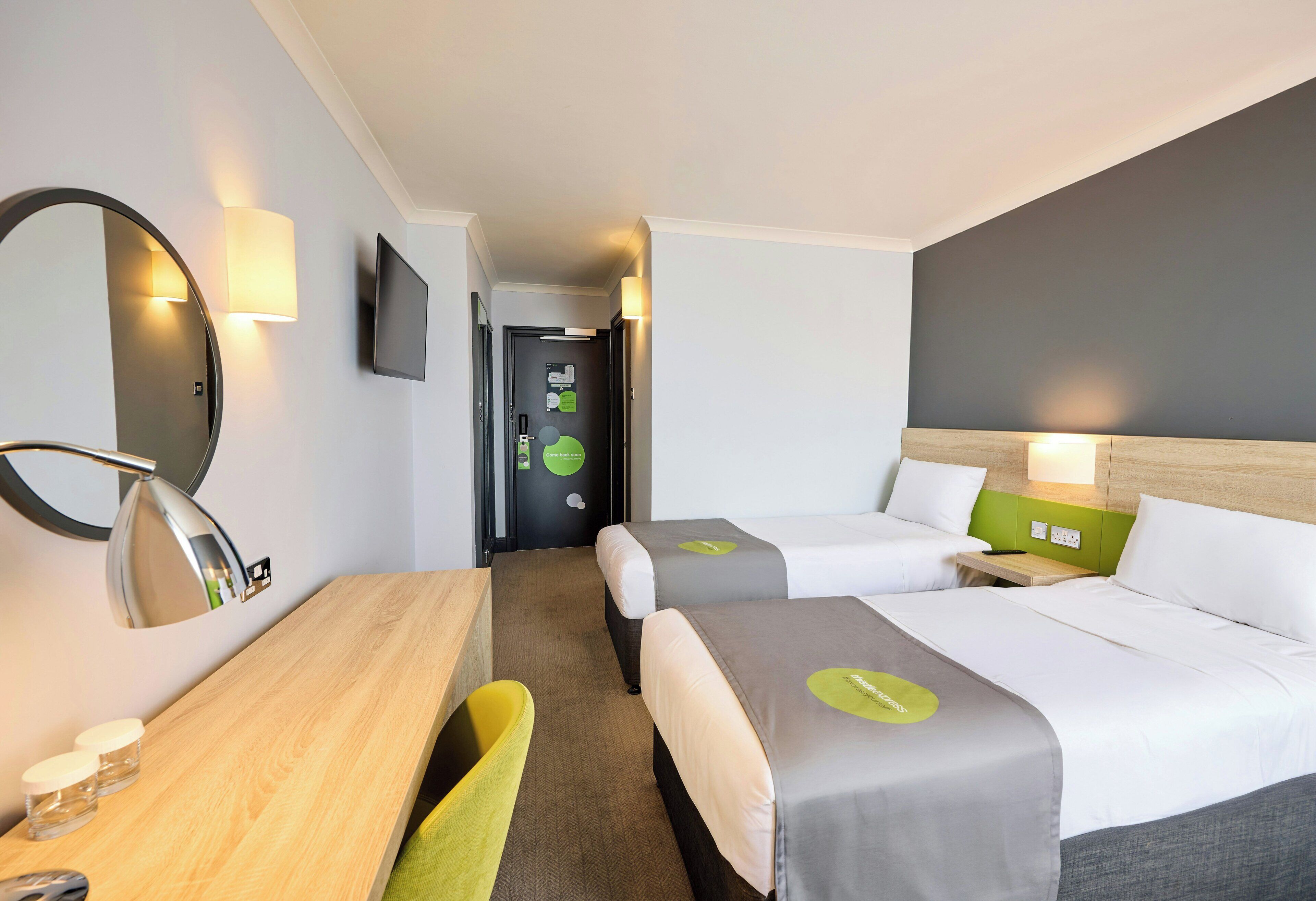A hotel room with two beds, a desk, a mirror, and a television.