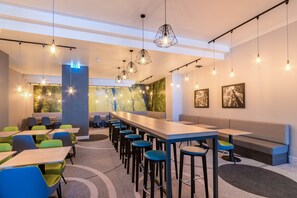 A modern lounge area with a long table, multiple chairs, and bar stools. There are hanging light fixtures and framed pictures on the walls.