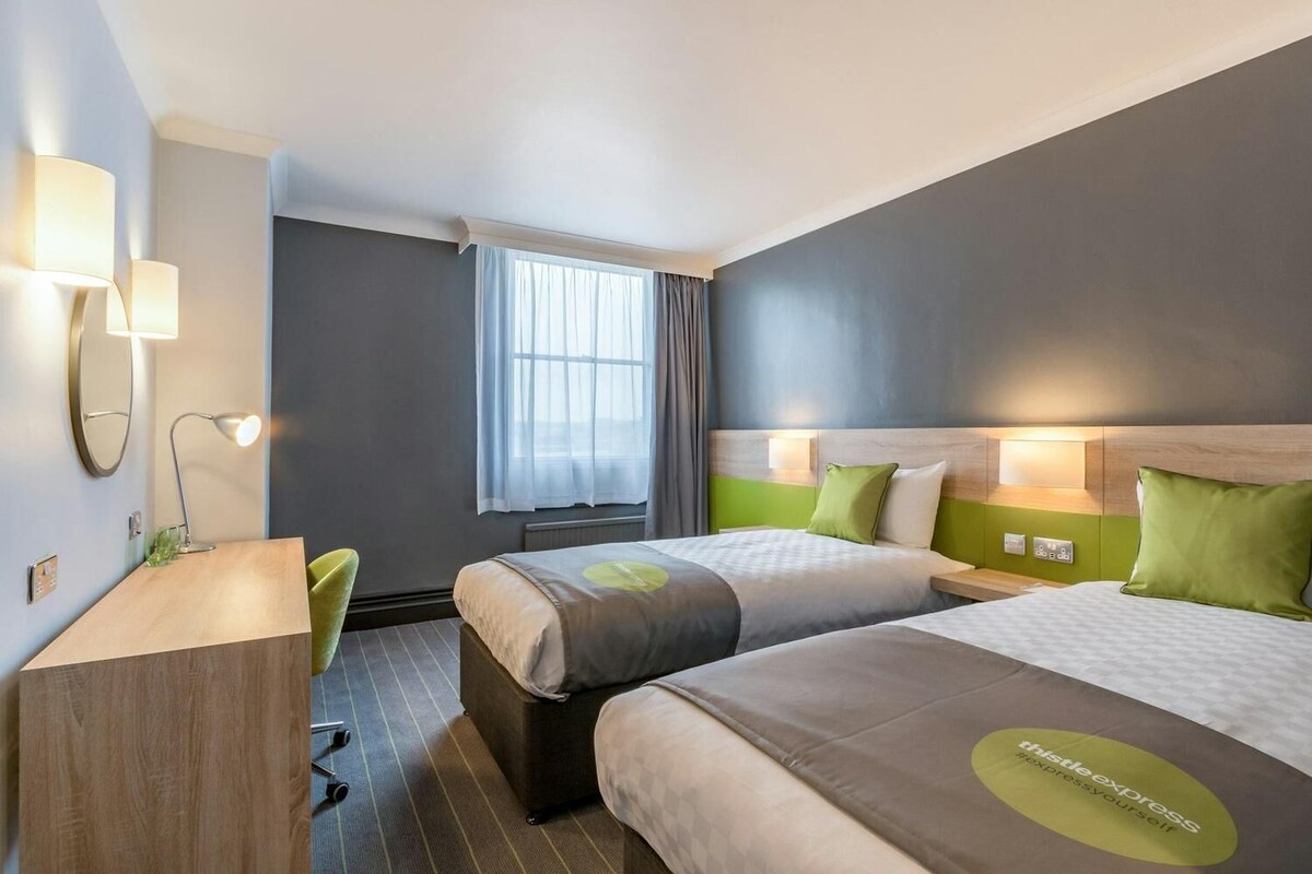 A hotel room with two beds, a desk, a chair, a window with curtains, and wall-mounted lamps.