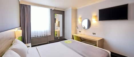 A hotel room with a bed, a bedside table with a lamp, a mirror, a TV mounted on the wall, and a window with curtains.
