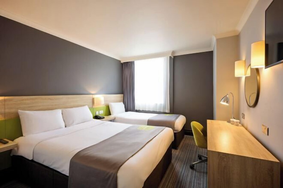 A hotel room with two beds, a desk, a chair, a nightstand, a lamp, and a large window with curtains.