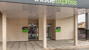 L’entrée d’un bâtiment Thistle Express, caractérisée par une façade en verre transparent, un sol carrelé et une porte d’entrée bien visible.