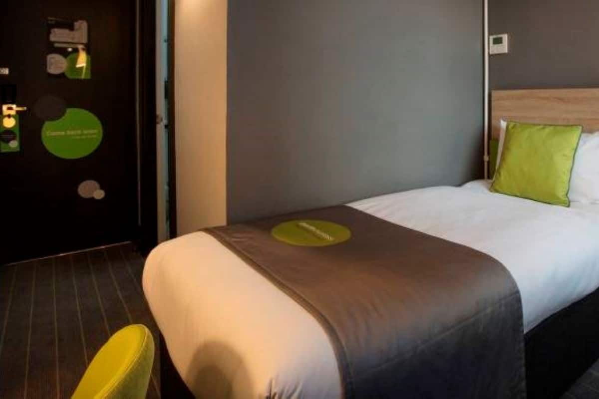 A hotel room with a bed, a chair, and a door with a green circular sign.