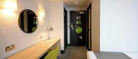 A hotel room with a wooden desk, a green chair, a bed, and a door with a 'Come back soon' sign.