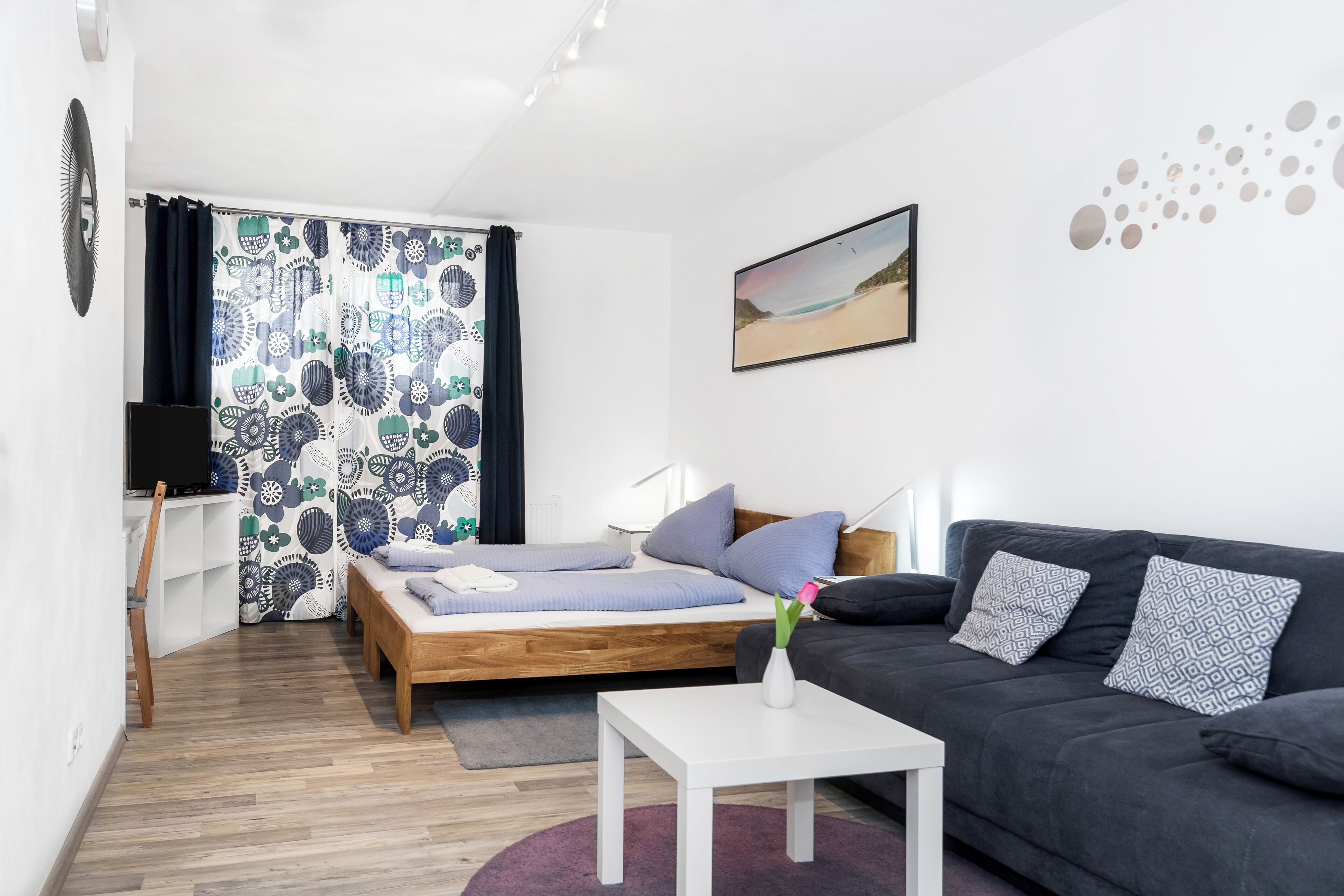Una habitación moderna con una cama, un sofá, una mesita, un televisor y una ventana con cortinas estampadas.
