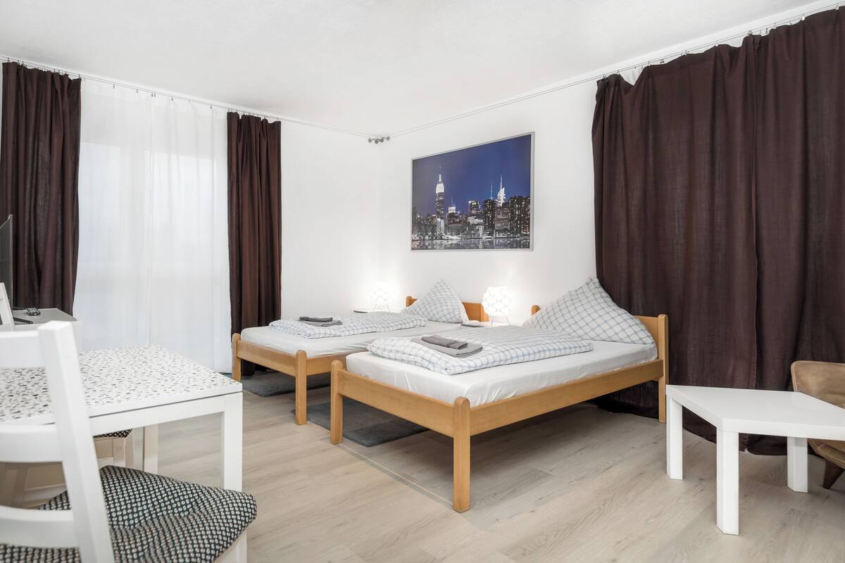 A modern hotel room with two single beds, a small table, a chair, and a cityscape painting on the wall.