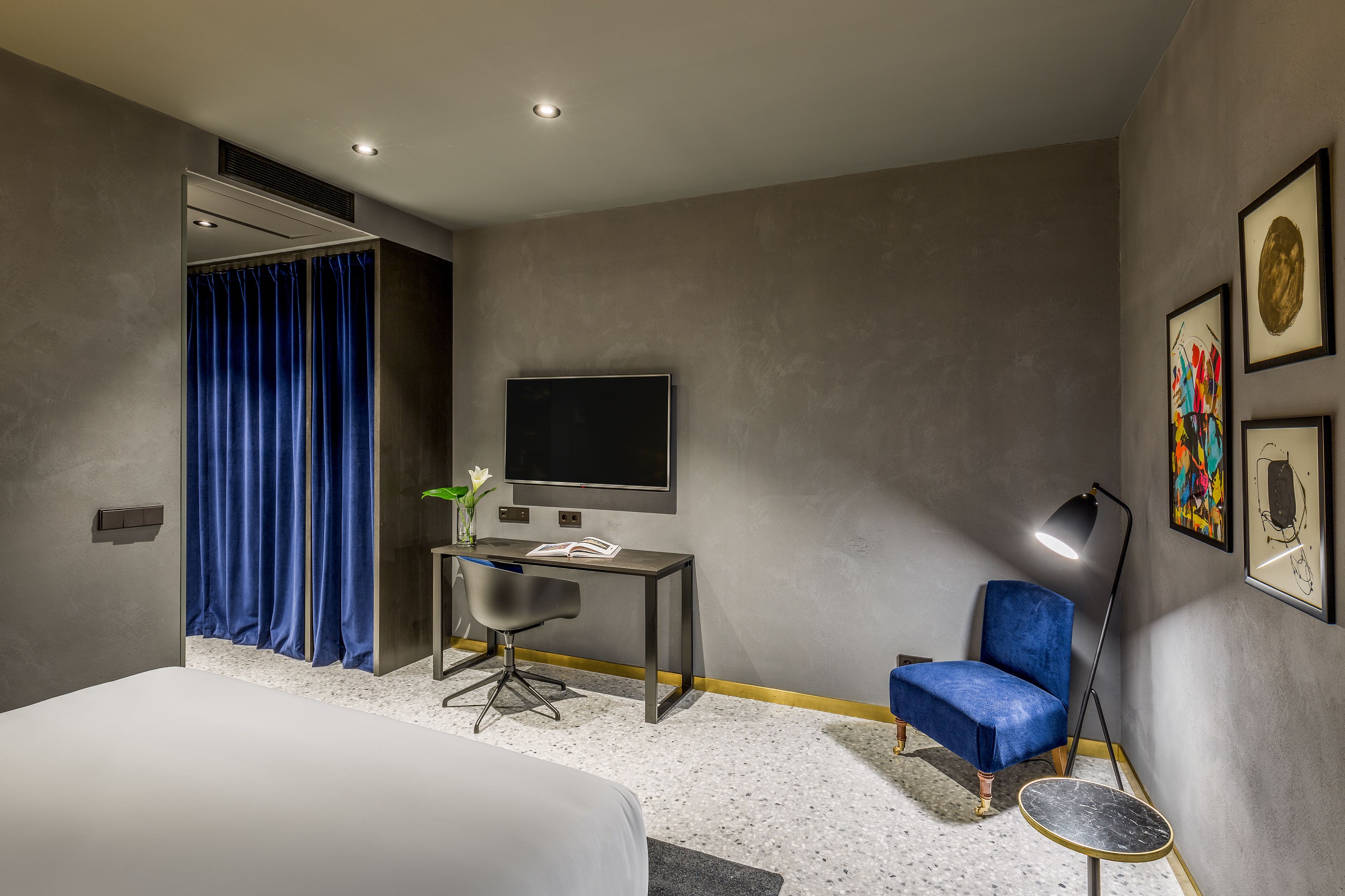 A modern hotel room with a bed, a desk with a chair, a TV, a blue armchair, and artwork on the walls.