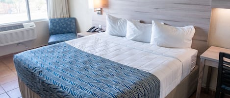 Habitación de hotel con una cama grande, una colcha azul y blanca, un cabecero de madera, un cuadro de un océano azul en la pared, una silla, una mesita de noche con una lámpara y una ventana con cortinas.