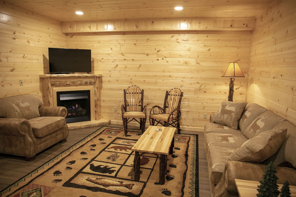 A log cabin interior with a fireplace, leather sofas, wooden chairs, a TV, and a lamp.