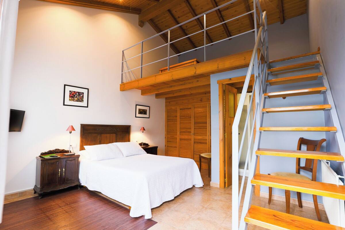 A bedroom with a wooden bed, a nightstand, a desk, a chair, a ladder, and a wooden door.