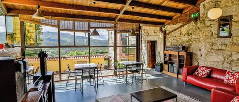 A spacious living area with a stone wall, a red leather sofa, a dining table with chairs, and a view of mountains through large windows.
