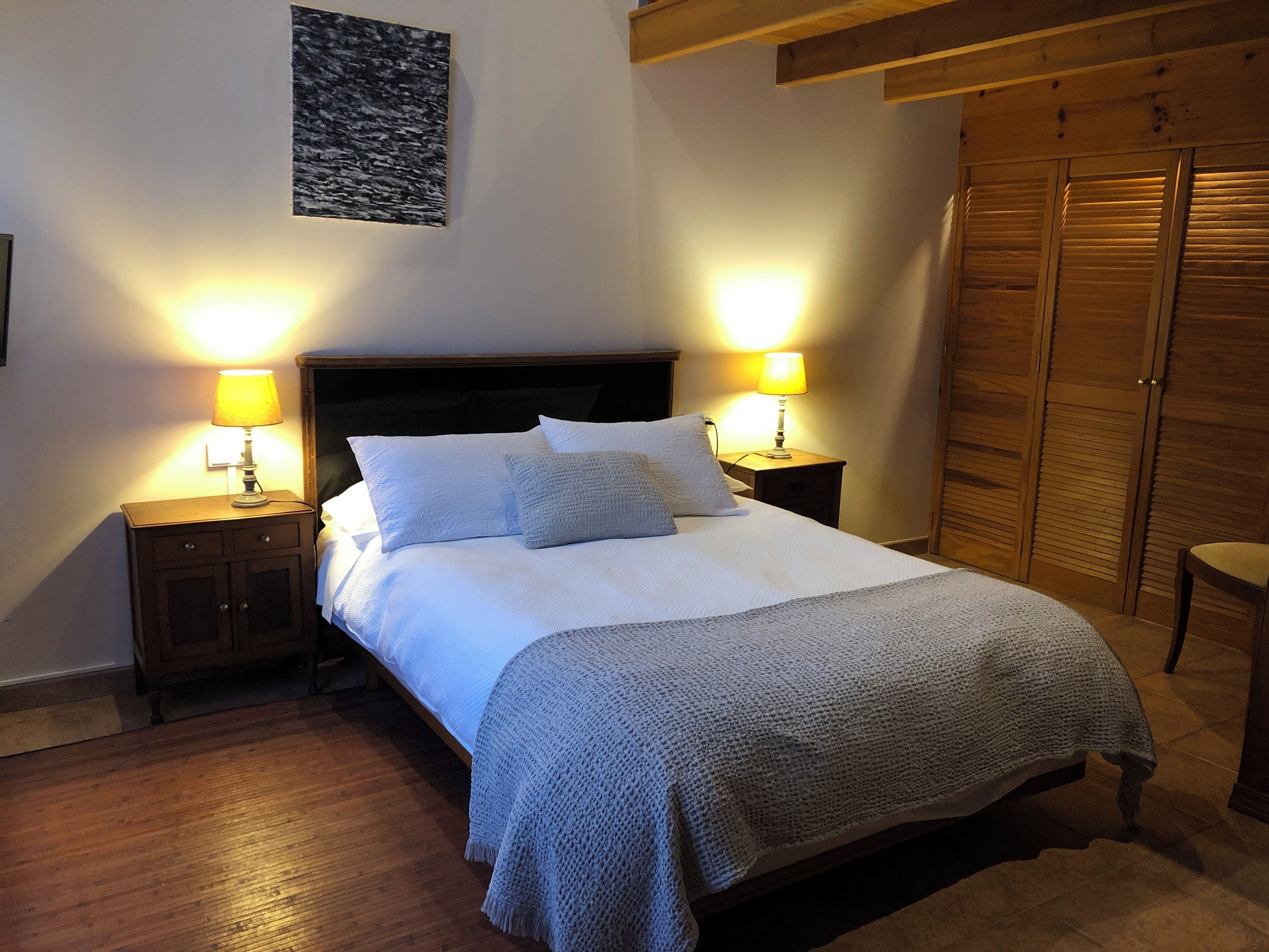 Una cama bien hecha con edredón blanco y dos almohadas, flanqueada por dos mesitas de noche con lámparas, un cabecero de madera, un armario de madera y un cuadro enmarcado en la pared.