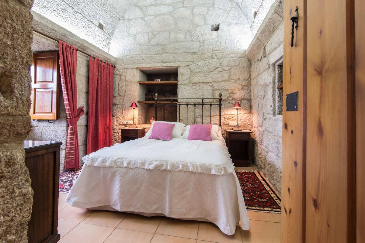 A bedroom with a stone wall, a wooden bed, a red curtain, and a nightstand with a lamp.