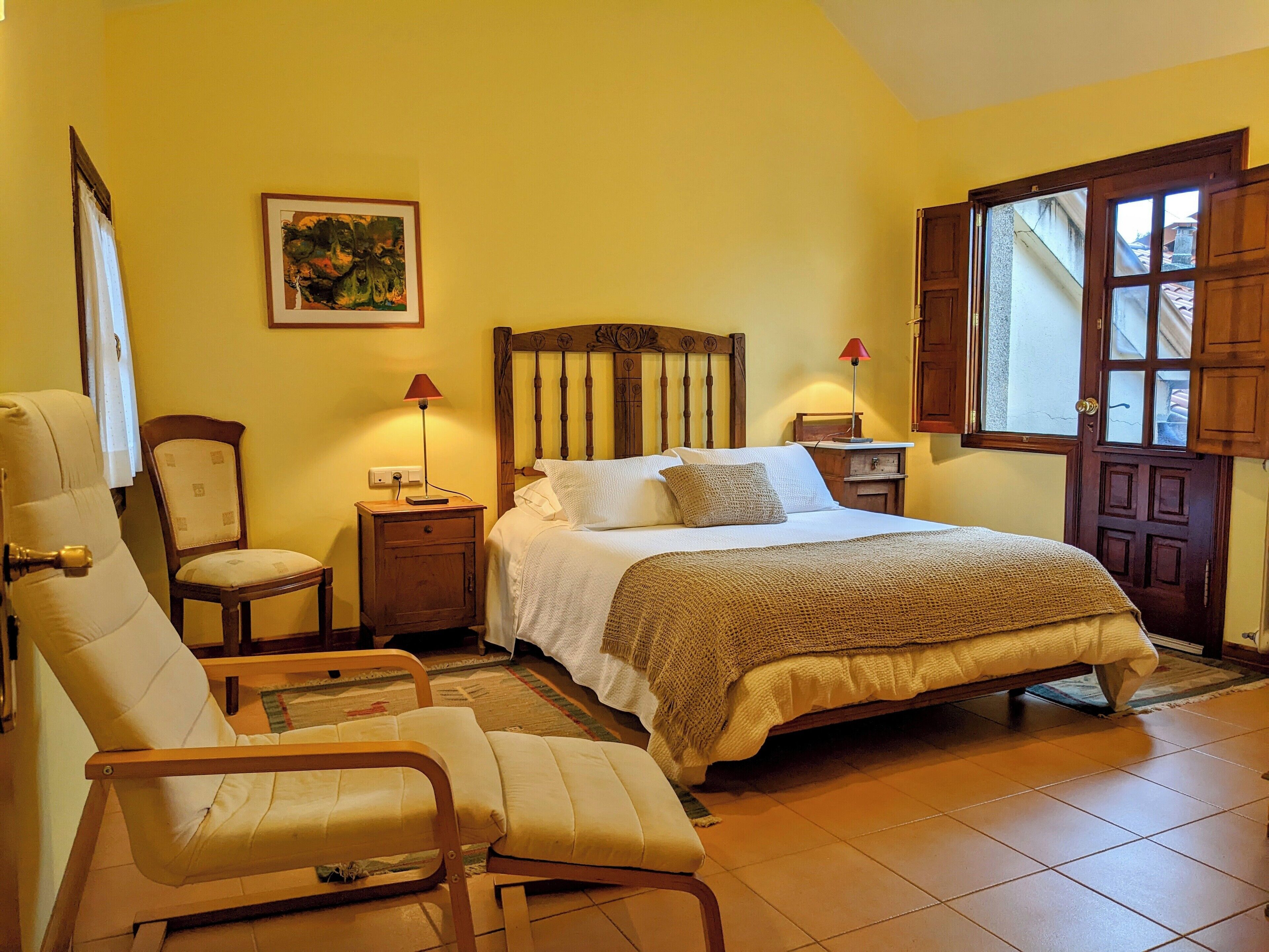 Un dormitorio con cama, dos mesitas de noche, una silla, una lámpara, un cuadro y una ventana con contraventanas de madera.