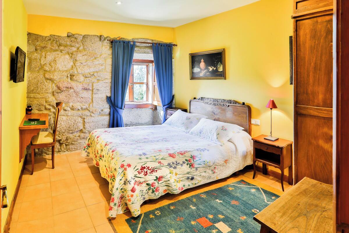A bedroom with a stone wall, a bed with floral bedding, a wooden nightstand, a chair, and a small table.