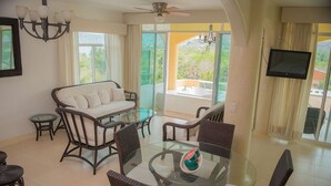 A modern living room with a glass dining table, wicker furniture, a sofa, and a large window offering a view of greenery.