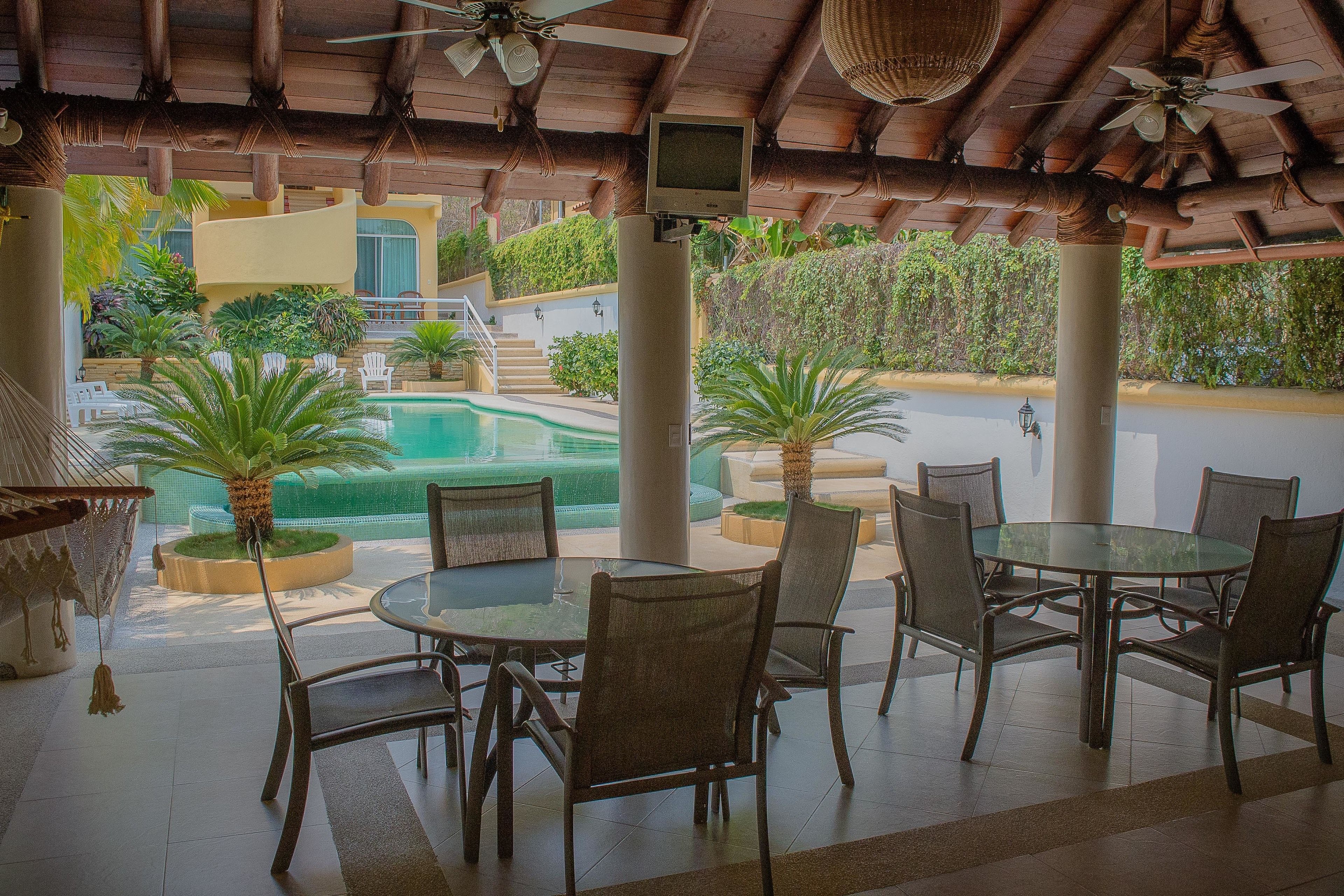 A covered outdoor dining area with a pool view, featuring a round glass table and chairs, wooden ceiling, and a TV screen.