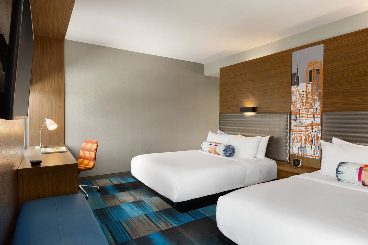 A hotel room with two beds, a desk with a lamp, a chair, and a large abstract artwork on the wall.