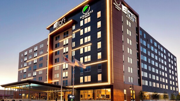 A modern hotel building with multiple floors, a prominent entrance with a covered area, and illuminated signage. - Aloft by Marriott Dallas Love Field (Dallas)