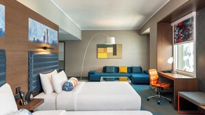 Et moderne hotelværelse med seng, skrivebord, stol og sofa. Et bybillede hænger på væggen, og der er en lampe på skrivebordet.