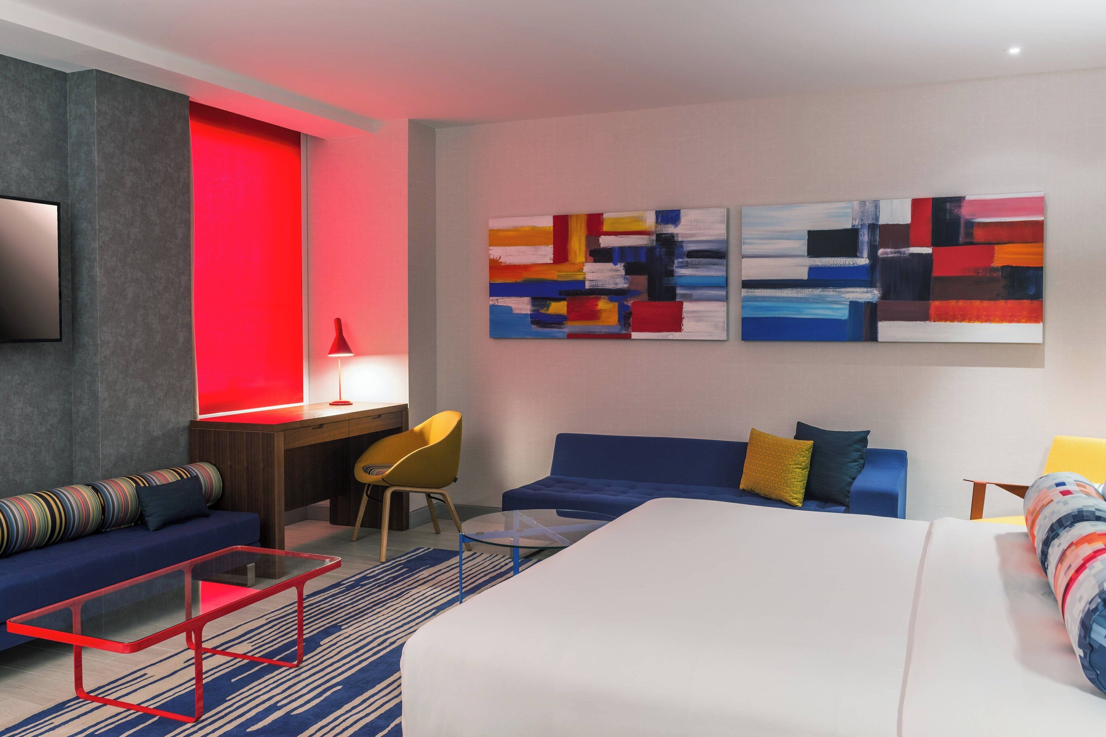 Ein modernes Hotelzimmer mit einem großen Bett, einer Sitzecke, einem Schreibtisch mit Stuhl, einem Fernseher und abstrakter Kunst an der Wand.