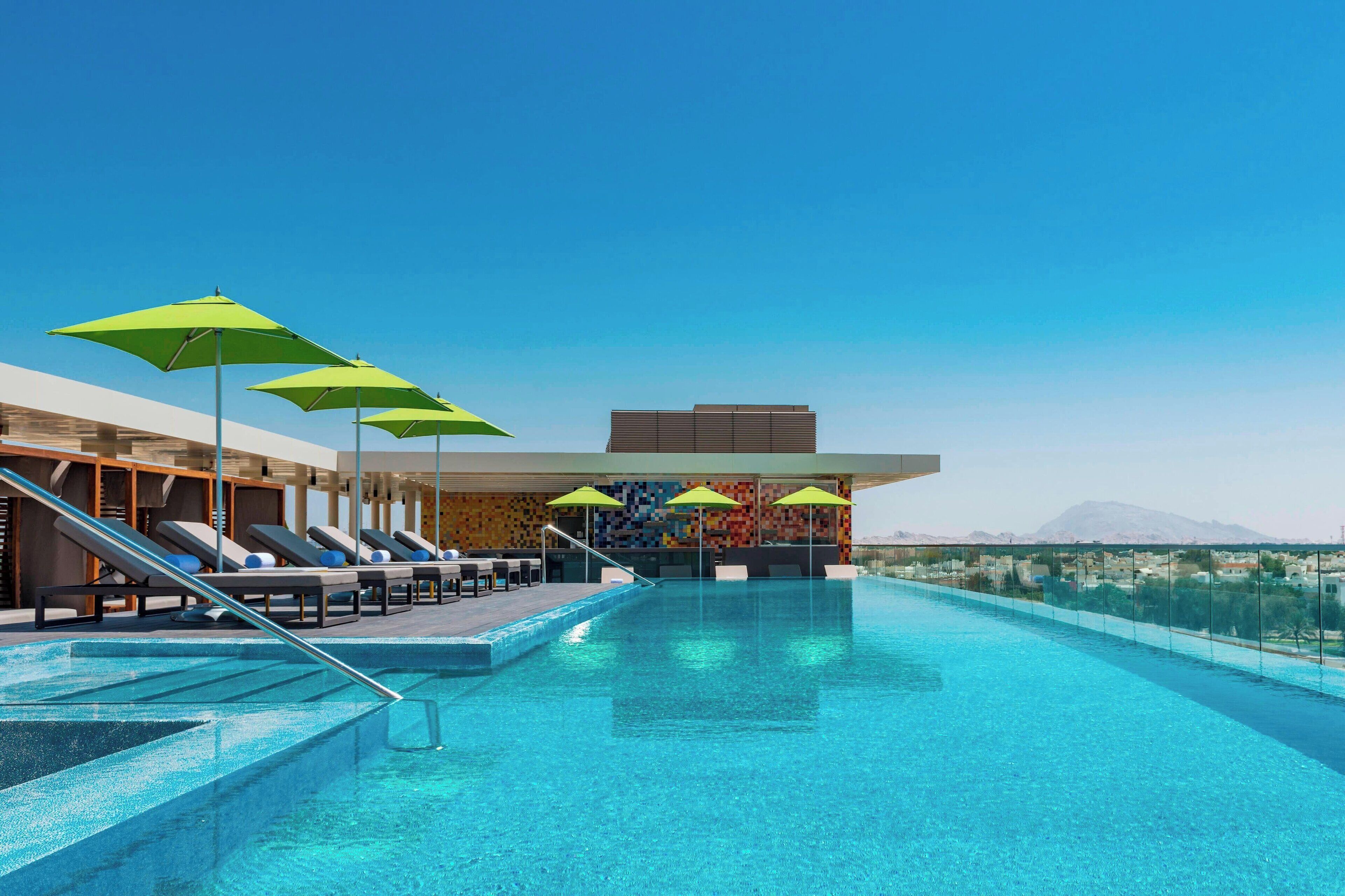 Un área de piscina moderna con sillones de descanso y sombrillas, un cielo azul despejado y vistas a montañas al fondo.
