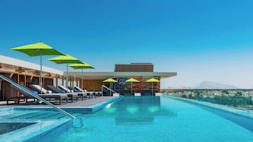 Un área de piscina moderna con sillones de descanso y sombrillas, un cielo azul despejado y vistas a montañas al fondo.