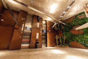 Un ascensor moderno con pantalla digital, una pared de madera con diseño geométrico y una pared verde con plantas.