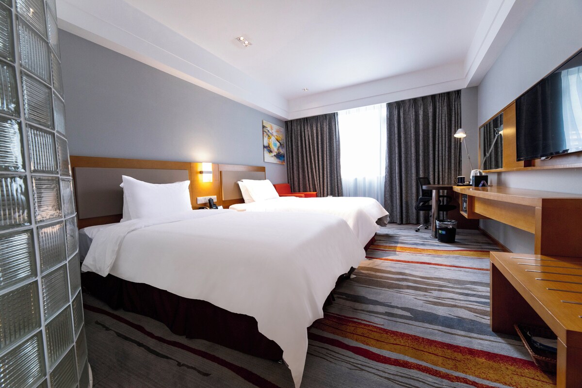 A hotel room with two beds, a desk, a television, and a large window with curtains.