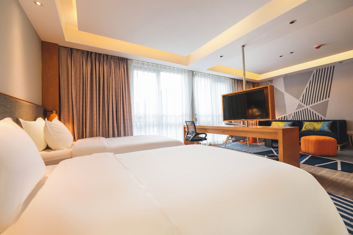 A hotel room with two beds, a desk, a television, and a large window with curtains.