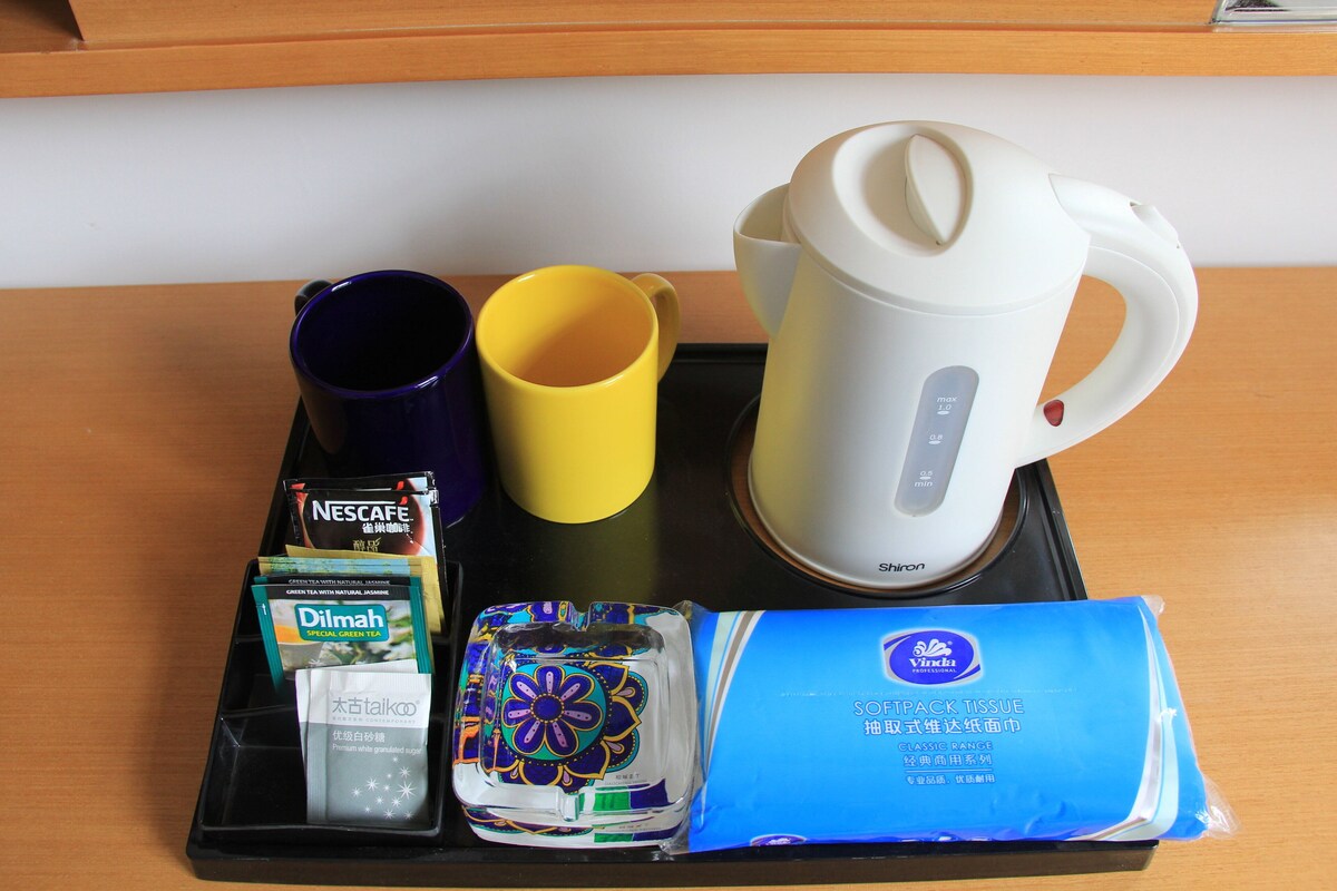 A tray with a white electric kettle, a purple and a yellow mug, a Nescafe coffee packet, a Dilmah tea packet, a pack of Vinda tissue, and a decorative item.