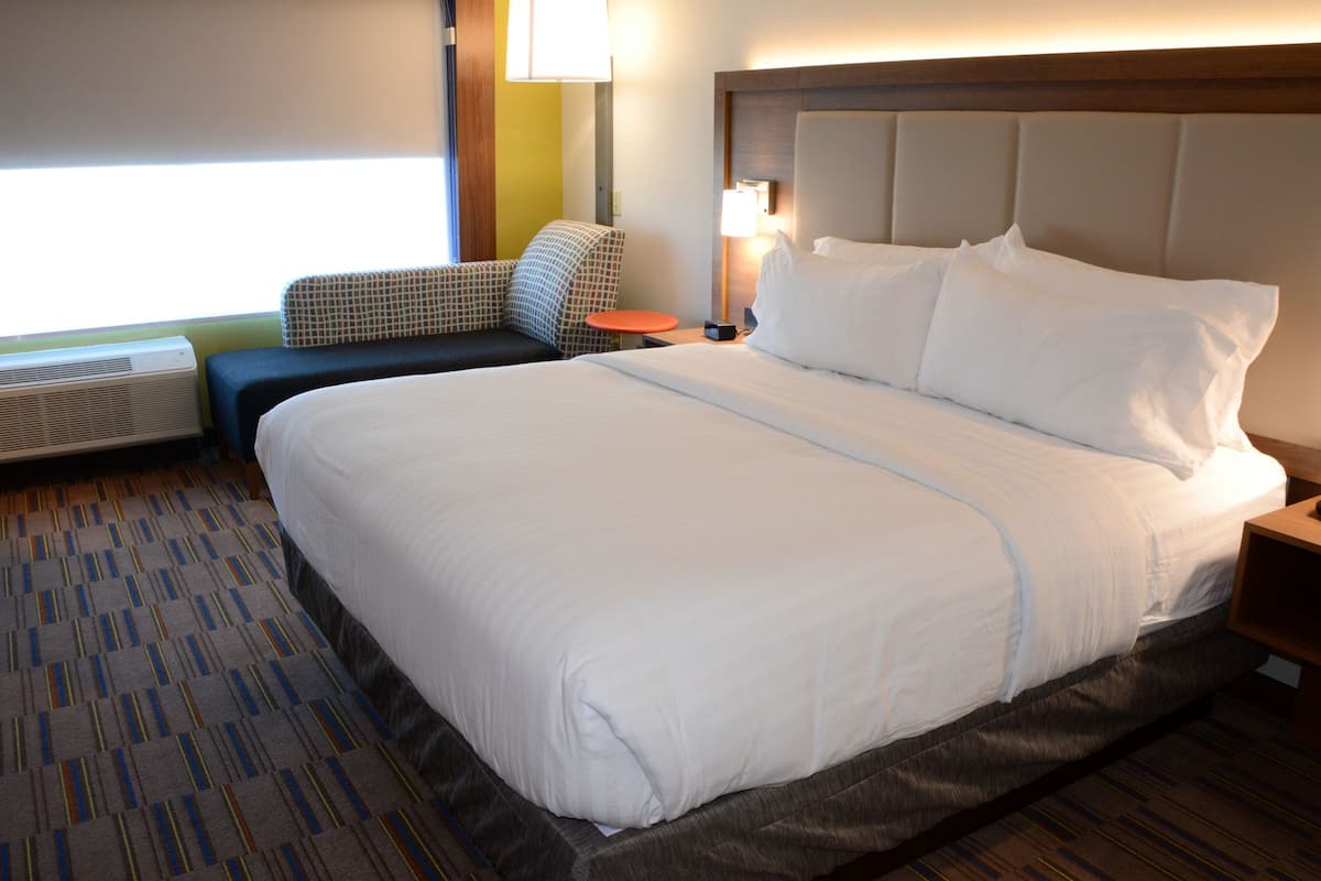 A hotel room with a large bed, a small table, a chair, and a window with curtains.