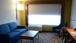 A hotel room with a blue sofa, a small wooden table, a desk with a chair, a standing lamp, and a large window with blinds.