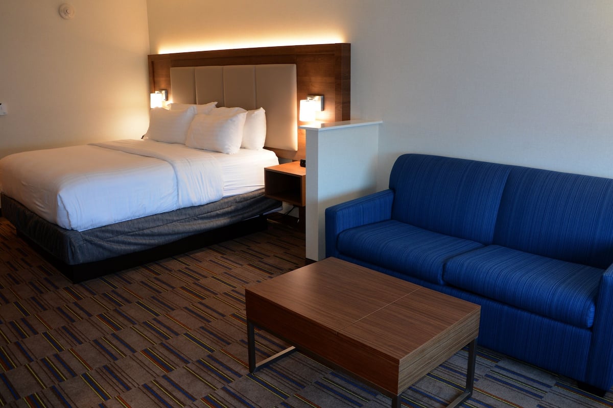 A hotel room with a bed, a blue sofa, a wooden coffee table, and a nightstand with a lamp.