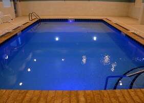 Una piscina interior rectangular con un tono azulado, bordes de azulejos y una escalera de metal.