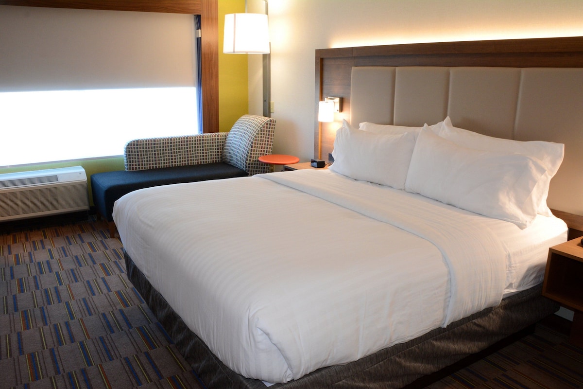 A hotel room with a large bed, a small table, a chair, a heater, and a window.