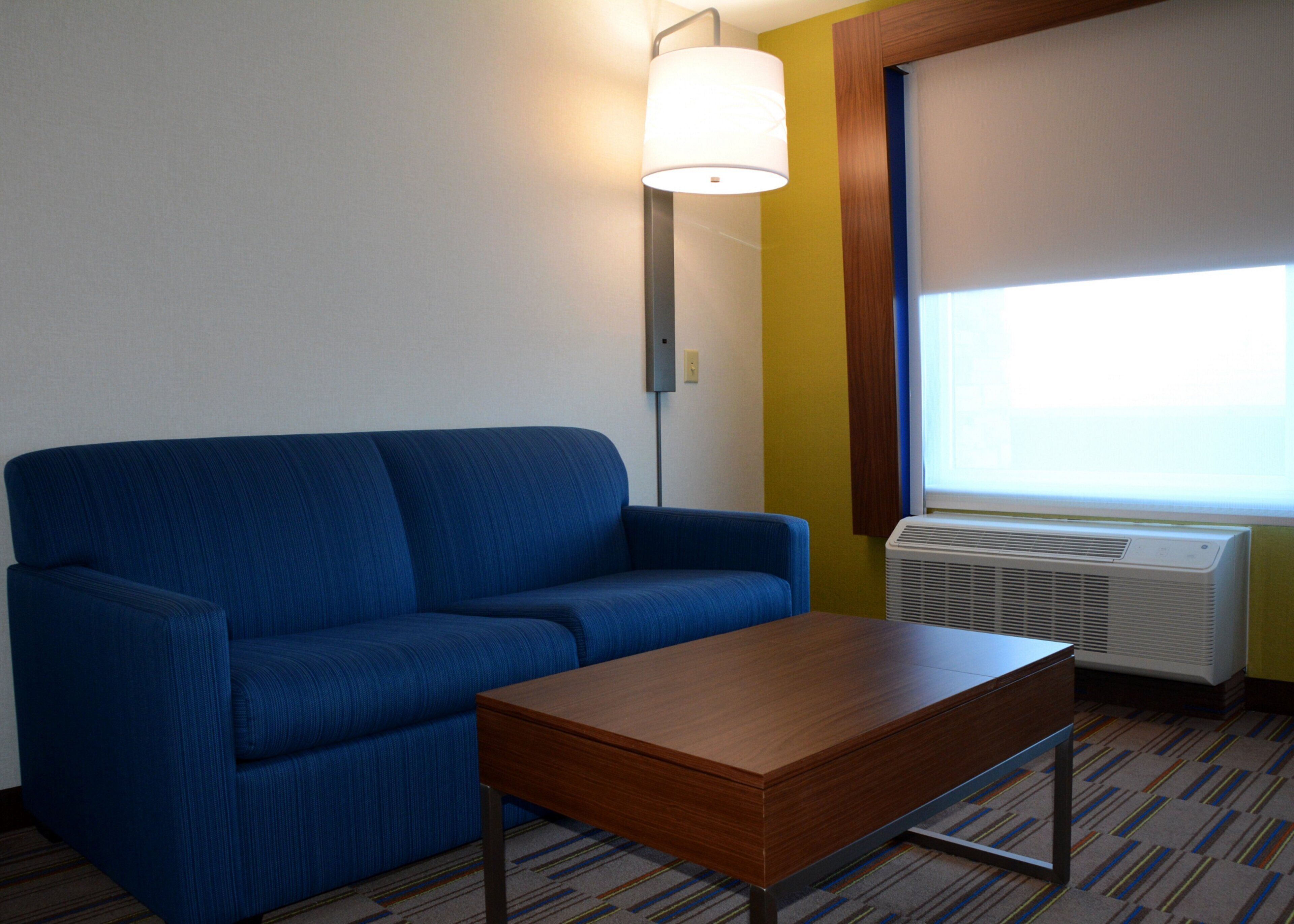 A room with a blue sofa, a wooden table, a lamp, an air conditioner, and a window with blinds.