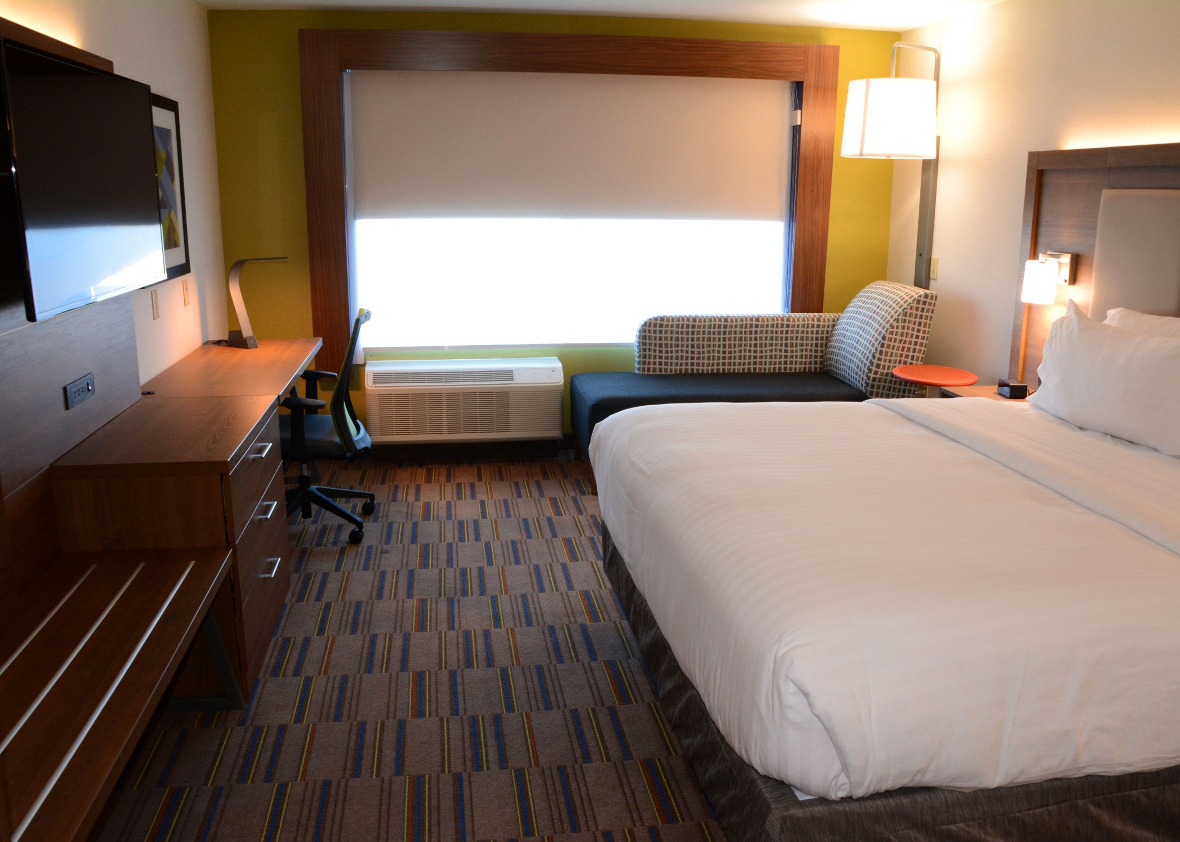 A hotel room with a large bed, a desk with a chair, a flat-screen TV, a window with blinds, and a lamp.